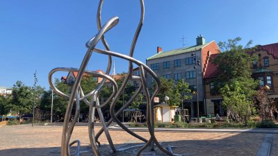 Konstverket Omväg i slingrande stålformation i förgrunden på gult marktegel. Glasskiosk, grönska och flerbostadshus i bakgrunden.