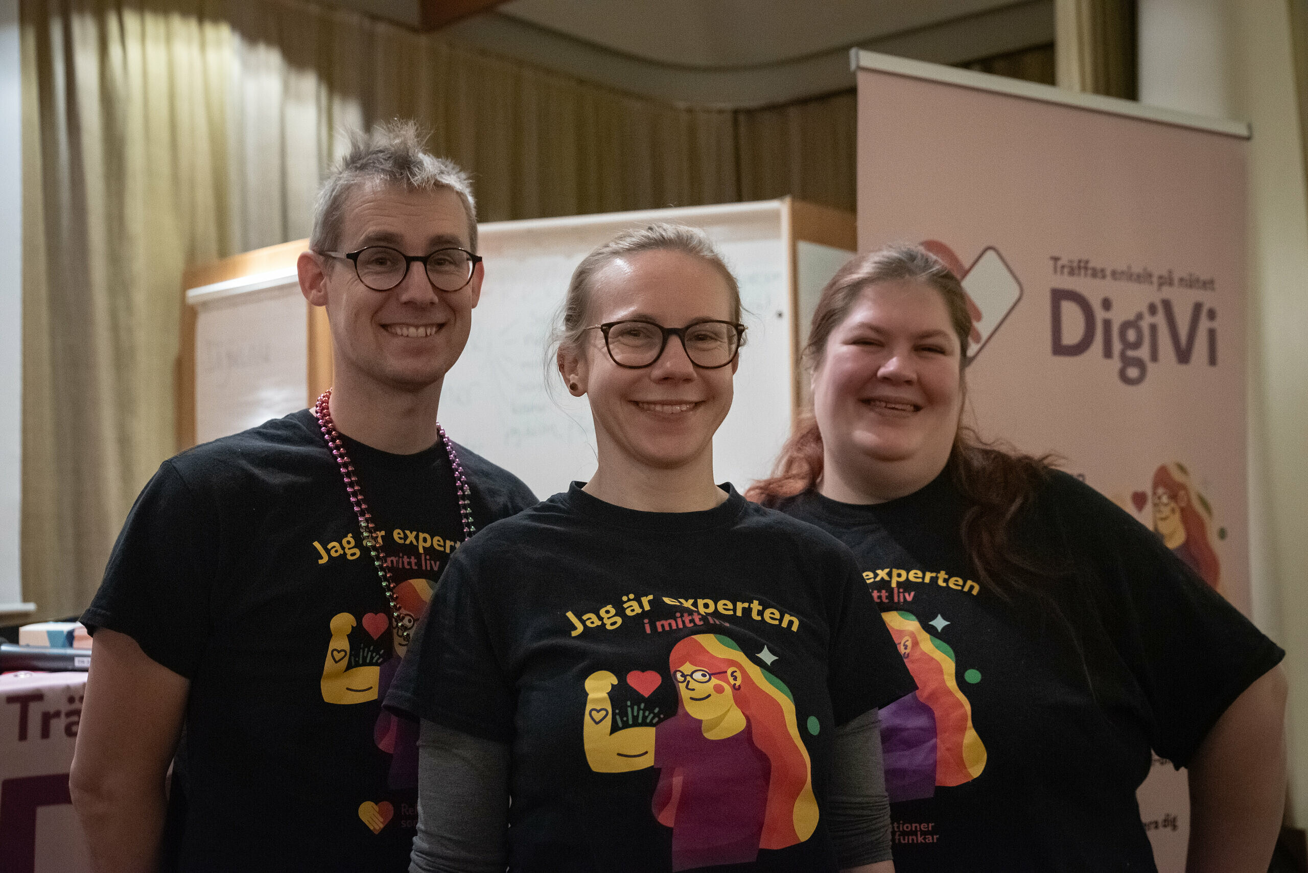 En man och två kvinnor står bredvid varandra och tittar på kameran. De alla har på sig samma t-shirt som visar illustration på en person med håret regnbågsfärgat. Ovanför illustrationen står texten: "jag är experten i mitt liv".