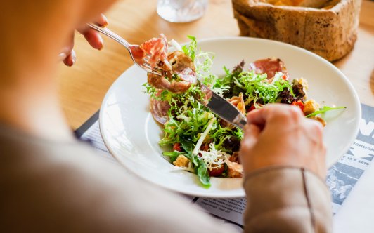 Tallrik med sallad och annat. I förgrunden en person som man bara ser händerna på när de håller i bestick.