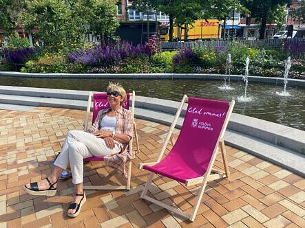 Två rosa solstolar framför dammaen. Frodig växklighet bakom dammen. I en av solarna sitter en blond kvinna i ljusa kläder och med solglasögon.
