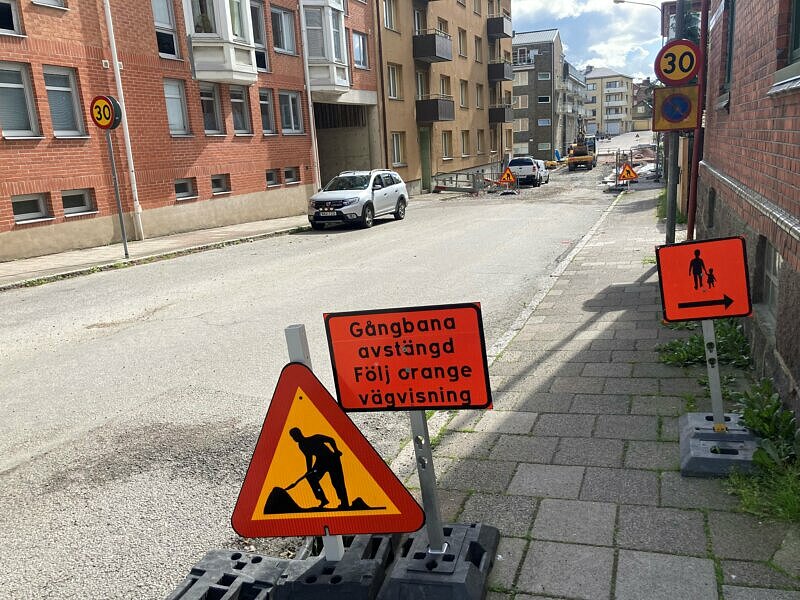 Varningstriangel och röd skylt med text att gatan är avstängd. Gata med parkerade bilar och tegelhus bakom.